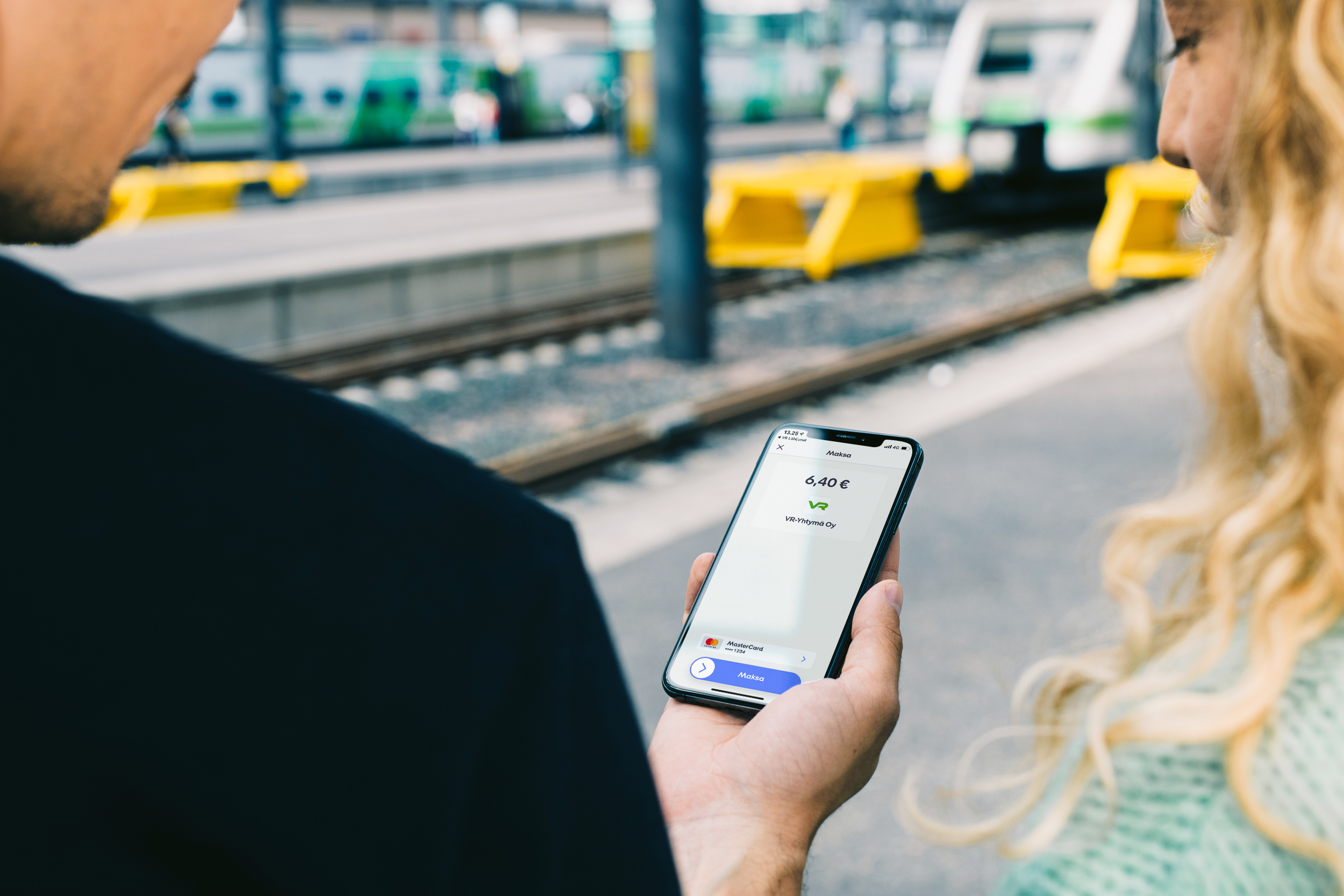 Mies ja nainen rautatieasemalla, mies puhelin kädessä maksamassa junalippua MobilePaylla